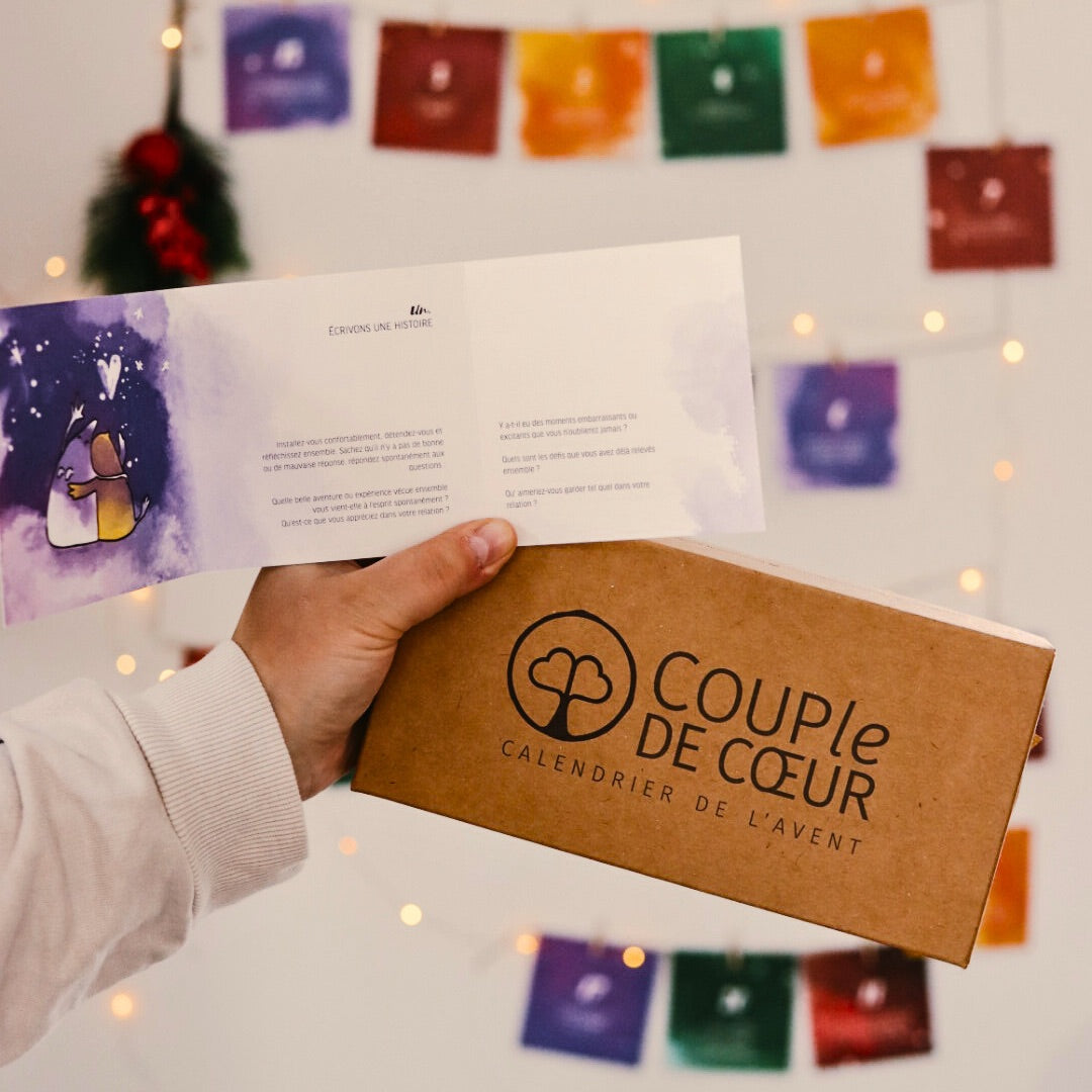 Main tenant le calendrier de l'Avent COUPle DE CŒUR édition 1 et une carte ouverte, devant une guirlande de cartes