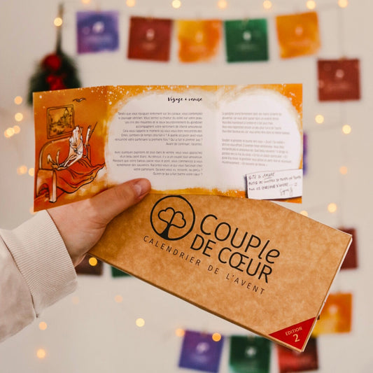Main tenant le calendrier de l'Avent COUPle DE CŒUR édition 2 et une carte ouverte, devant une guirlande de cartes